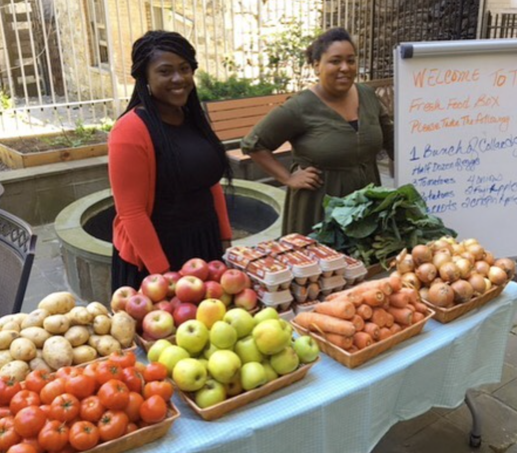 Two smiling women stand behind a table laden with fresh produce, including tomatoes, potatoes, apples, eggs, carrots, onions, and leafy greens, likely at a farmers market or a community food distribution event, with a whiteboard displaying information about a "Fresh Food Box."